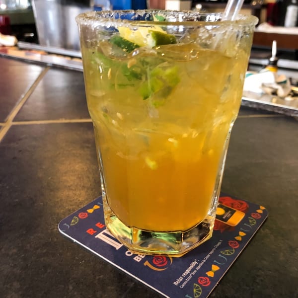 Yellow cocktail with lime, mint, and a salt rim on a coaster at a bar.
