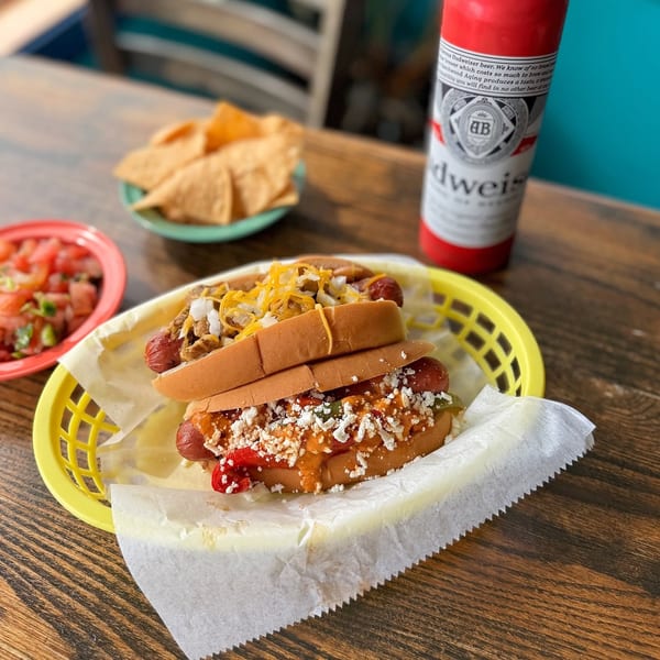 Two loaded hot dogs in a basket with chips, salsa, and a Budweiser on a wooden table.