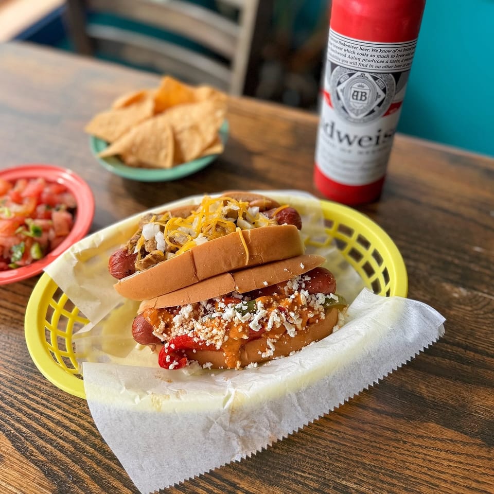 Two loaded hot dogs in a basket with chips, salsa, and a Budweiser on a wooden table.