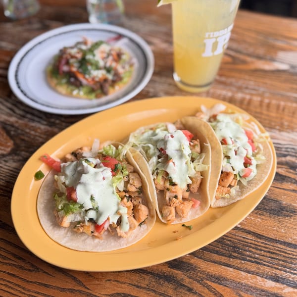 Three chicken tacos with lettuce, tomato, and green sauce on a yellow plate, with a tostada and drink.