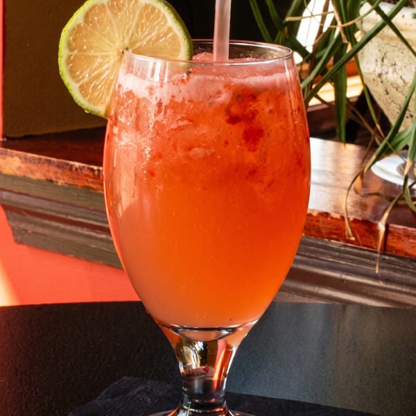 Vibrant red blended cocktail with a lime slice and straw on a dark table, with plants in the background.