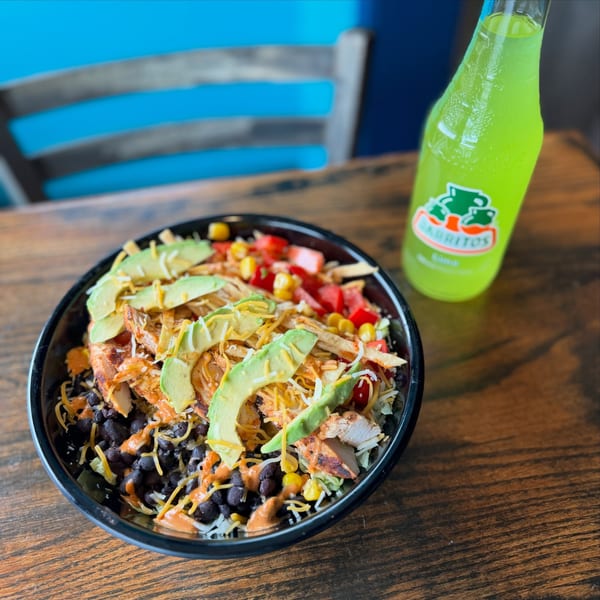 Chicken bowl with avocado, black beans, corn, cheese, and a green Jarritos soda on a wooden table.