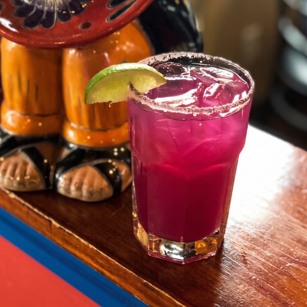 Vibrant pink cocktail with ice, a salted rim, and lime wedge on a wooden bar next to a colorful ceramic figurine.