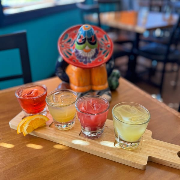 Four vibrant drinks on a wooden flight board with orange slices and a colorful sombrero figure.