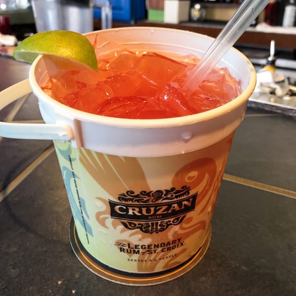 Large red rum punch in a Cruzan Rum bucket with a lime wedge and straws on a bar.